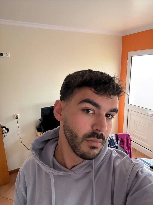 A young man with short dark hair and a beard, wearing a gray hoodie, takes a casual selfie in a room with white and orange walls. The image pairs with a "Good Morning" post requesting users to drop their $SOL (Solana) cryptocurrency addresses, likely to send tips or airdrops.