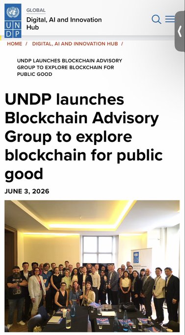 The image shows a group of people posing for a photo, with some standing near a window and others gathered around a table. There are at least 13 visible individuals in the picture. The text "UNDP Launches Blockchain Advice Group to explore blockchain for public good" is displayed on the screen above the image. This information suggests that the image might be related to an event or gathering organized by the United Nations Development Programme (UNDP), which aims to promote sustainable development and improve people's lives around the world.