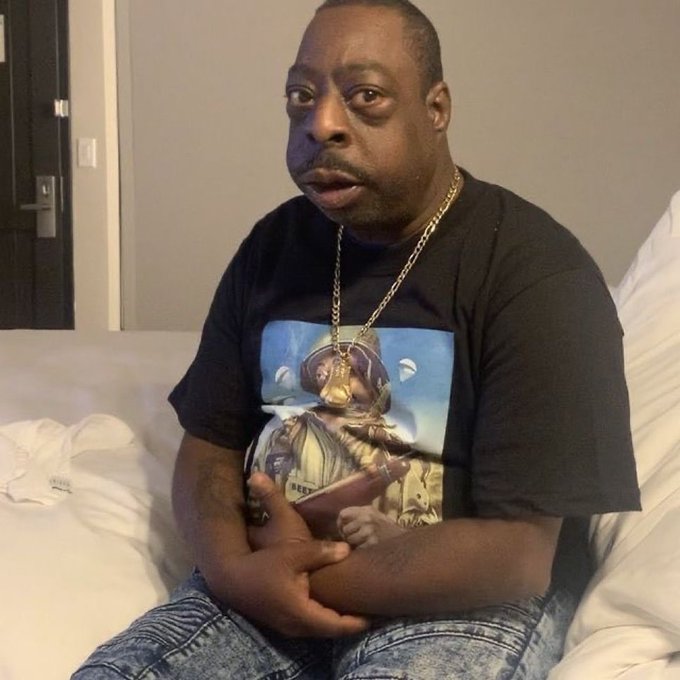 A photograph of a man with a mustache wearing a black graphic t-shirt featuring a robot design and a gold chain, sitting on a white bed and looking directly at the camera.