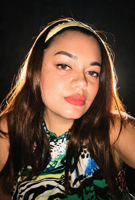 A selfie-style portrait of a woman with long brown hair, red lipstick, and a patterned top posing against a dark background.