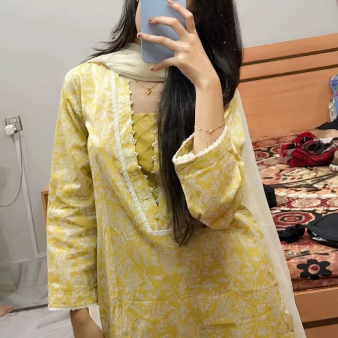 A woman takes a mirror selfie while wearing a yellow floral traditional South Asian outfit with a white dupatta, displaying delicate necklaces.