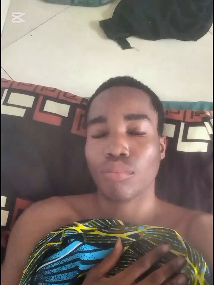 The image shows a man lying down on his stomach with his eyes closed, possibly sleeping or meditating. The background of the photo features a bedspread with a patterned design. There are also two bags visible in the scene - one near the top left corner and another towards the bottom right corner.
