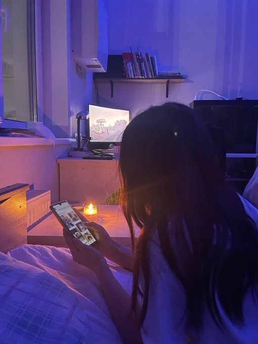 A woman with long dark hair sits in a dimly lit bedroom illuminated by blue and purple light, scrolling on her smartphone while a candle glows nearby.