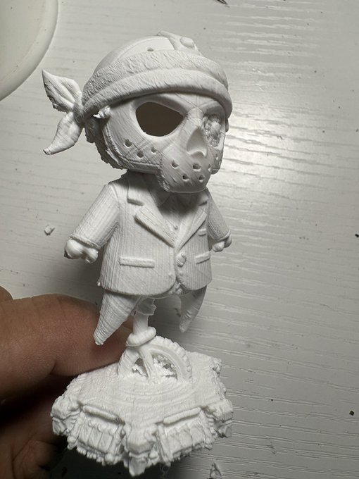 A small white 3D-printed figurine of a character wearing a hockey-style mask, bandana, and suit jacket, held between fingers over a work surface.