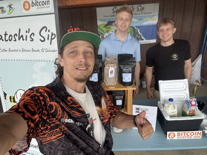 A man in a vibrant orange and black patterned shirt takes a selfie in front of 'Satoshi's Sip' coffee stand in El Salvador, with a Bitcoin Accepted Here sign visible.