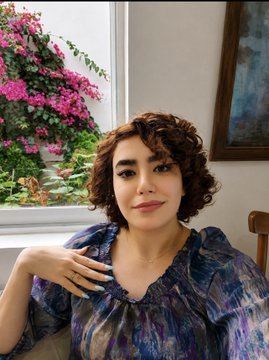 A woman with short curly brown hair and a nose ring wearing a blue patterned top poses for a portrait, with pink flowers in a window and framed artwork visible in the background.