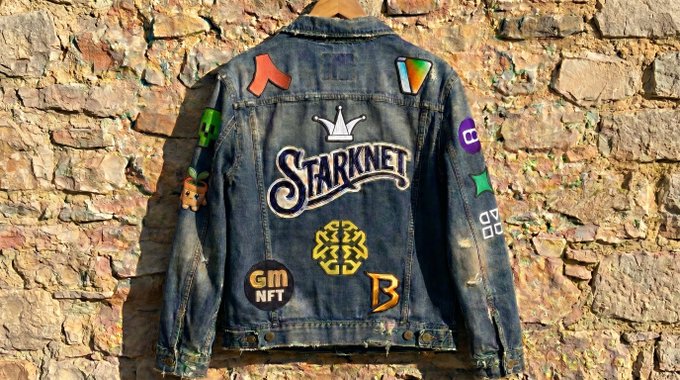 The image features a denim jacket hanging against a brick wall. The jacket has various patches and stickers on it, including one that reads "Starknet" and another with a logo for "Gm While researching L2s revenue". These patches and stickers add character and personalization to the jacket, making it unique and easily recognizable.