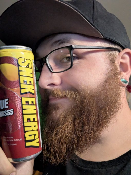 A bearded man with glasses and a black cap holding up a can of 'Snek Energy' Blue Raspberry energy drink.