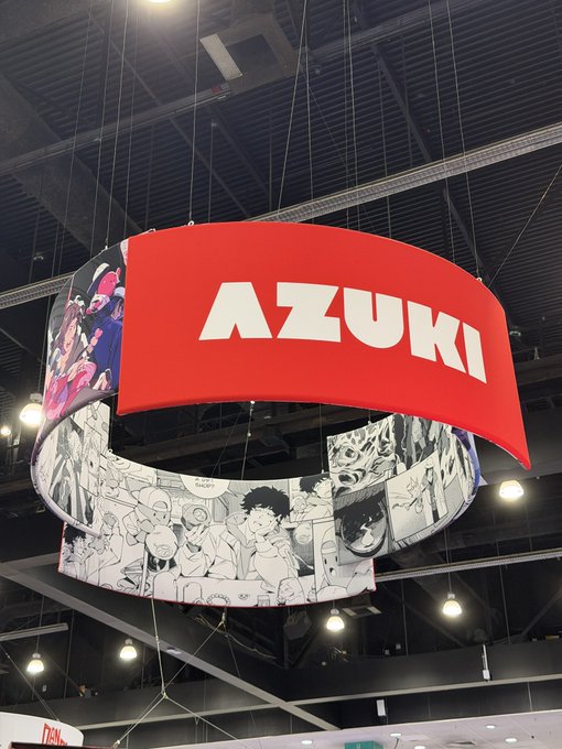 A large circular hanging banner displays the "AZUKI" logo in bold white letters on a red background, surrounded by black-and-white manga-style character artwork. The image appears to be from a convention or trade show booth promoting the Azuki brand.