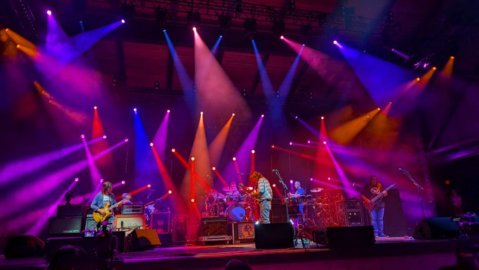 A live concert performance on a stage illuminated by vibrant pink, purple, blue, and orange spotlights, with a band playing guitars and drums.