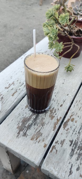 A tall glass of layered iced coffee with a white straw sitting on a weathered white wooden table, with a potted succulent plant in the background.