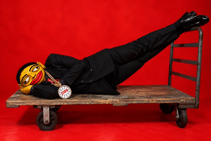 A figure in a black suit with a cartoonish Pepe mask, gold chains, and a "STONKS" clock medallion lies on a wooden cart against a red background.