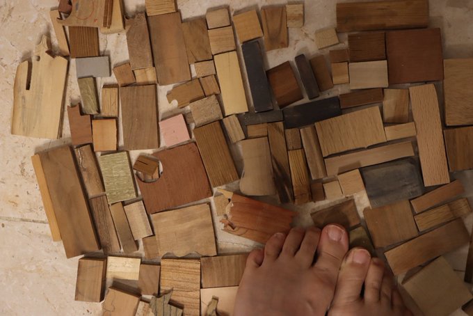 A collection of various scrap wood pieces in different shapes and brown tones scattered on a tiled floor, with a small foot visible at the bottom.