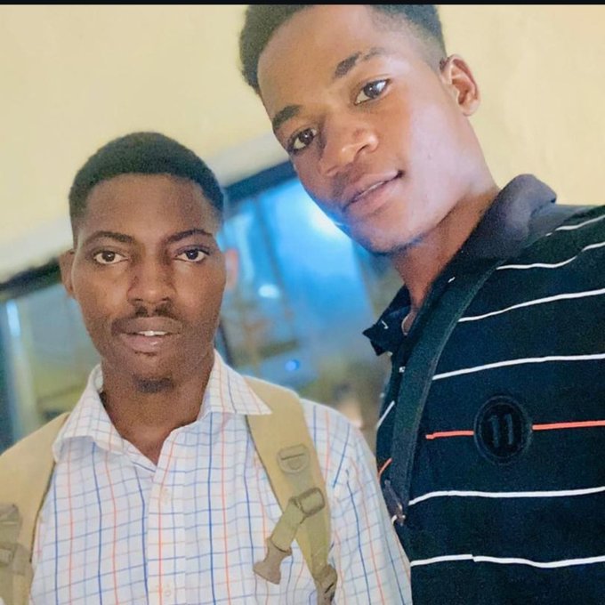 Two men posing together in a selfie, one wearing a checkered shirt with a backpack strap visible and the other in a black and white striped polo shirt.
