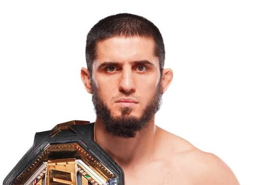 A portrait of MMA fighter Islam Makhachev, who has a beard and short hair, holding a UFC championship belt over his shoulder.