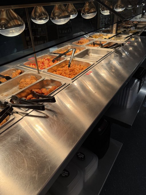 A nearly empty cafeteria buffet line with stainless steel food trays containing mostly picked-over chicken and pasta, with serving tongs and overhead heat lamps visible.