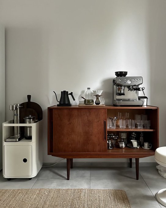 A mid-century modern wooden sideboard displays an espresso machine, pour-over coffee setup, and glassware, accompanied by a white rolling cart holding a grinder and kitchen accessories against a neutral wall. A woven jute rug lies on the gray tiled floor in the foreground.