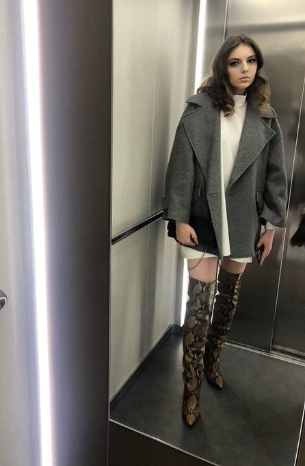 A woman standing in an elevator or metallic room, wearing an oversized gray coat, white dress, and snakeskin-patterned over-the-knee boots.