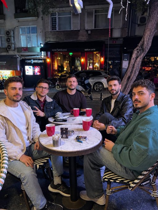 Five young men posing around a marble table with coffee mugs outside a café called "anya Tabiyası" (antre café) in what appears to be an evening street setting.