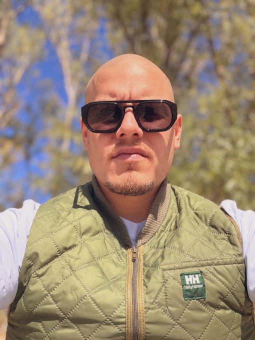 A man with a shaved head and goatee poses for a close-up selfie wearing dark sunglasses and a Helly Hansen (HH) quilted vest, likely moments after getting his head shaved. His freshly bald look ties into the post's announcement of losing his hair at Solana's Breakpoint event if he wins the Meme Lord Award.