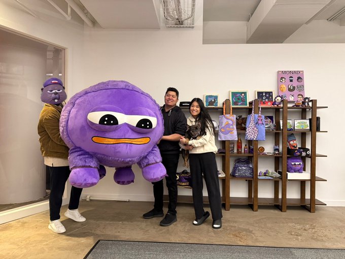 Two people pose with a small dog in what appears to be a Monad-themed office space, featuring a large purple mascot plush, branded merchandise, and shelves displaying "MONAD" posters and collectibles, celebrating their contribution award.