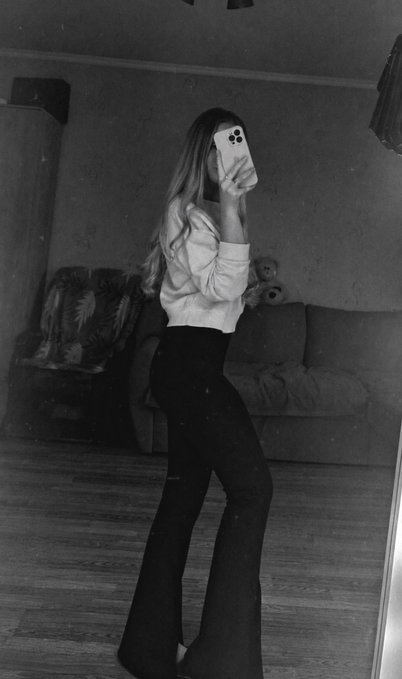 A black-and-white mirror selfie shows a young woman with long hair, dressed in a light blouse and dark flared pants, holding her phone up to capture the shot in a dimly lit living room. The moody, anonymous aesthetic complements the post's tone of emotional unavailability and guarded distance from new connections.