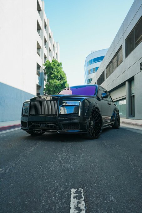 A black Rolls-Royce Cullinan SUV parked on an urban street between modern concrete buildings, with a pink and purple gradient roof visible in the background.