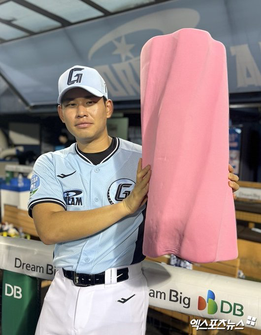 A baseball player in a KT Wiz uniform holding a large pink phallic-shaped object, photographed in a dugout with 'Dream Big' and 'DB' sponsor logos visible in the background.