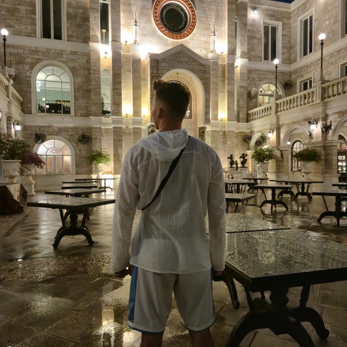 A young man viewed from behind, wearing a white hoodie and shorts, standing in a grand, European-style indoor courtyard or building lobby with stone columns, arched doorways, and tables.