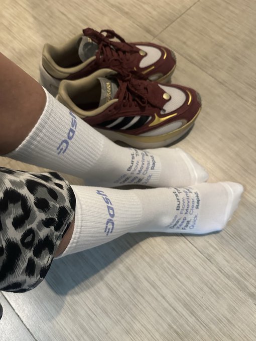 The image features a pair of white socks with blue text, one for each foot, placed next to a pair of red sneakers. The socks are positioned on a wooden floor, while the shoes are located near the left side of the frame. The caption "For when you really want to get there before anyone else." suggests that these items might be related to an event or activity where people often wear matching footwear and socks as part of their attire.