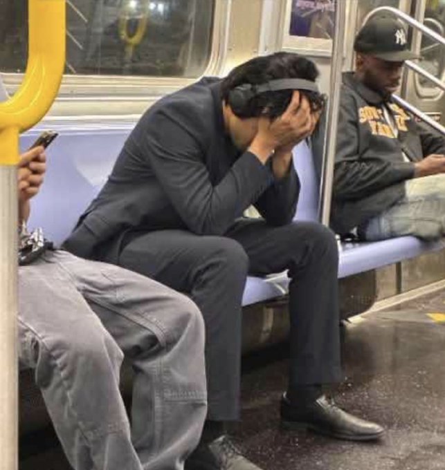 A man in a dark suit sits on a subway train with his head in his hands, wearing a large headset device, while other passengers sit nearby.