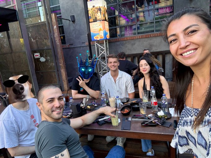 A group of friends enjoys a meal at an outdoor restaurant, with one person wearing a blue crown mask featuring green lights and a small dog with sunglasses perched nearby.