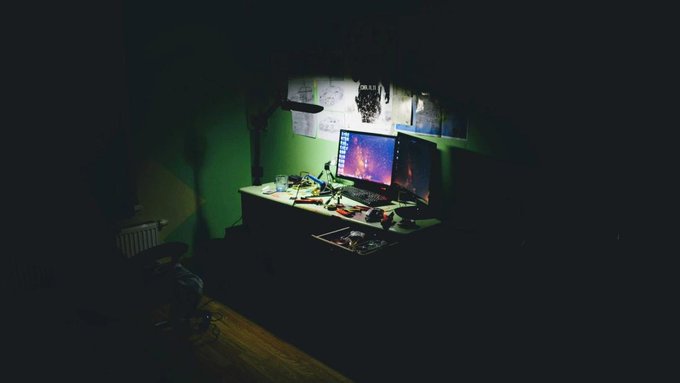 A shadowy, dimly lit desk setup with two glowing monitors displaying cosmic imagery and scattered papers, evoking the secretive, isolated environment of dark web operators working late into the night.