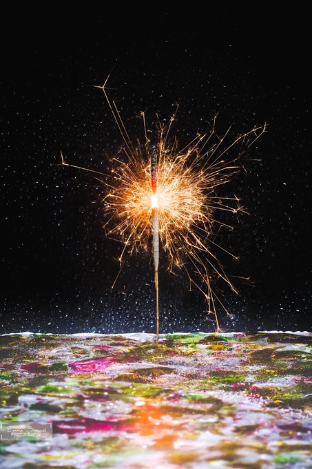 A sparkler burning brightly in the darkness, creating a burst of golden sparks that scatter across a black background above a colorful, textured surface.