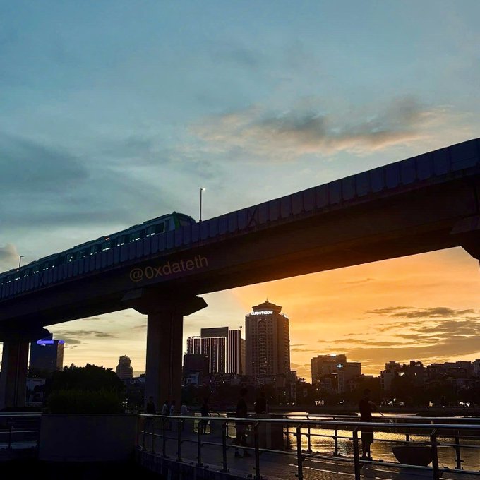 An elevated train crosses an overpass at sunset over a waterfront walkway, with the city skyline including the 'Luxview' building visible in the background. A watermark reading '@oxdateth' appears on the bridge.