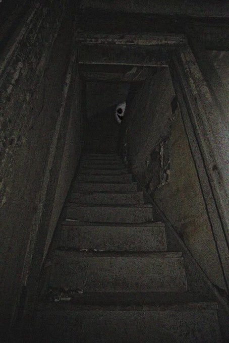A dark, eerie photograph taken from the bottom of a narrow staircase looking up toward a shadowy figure with glowing eyes standing at the top.