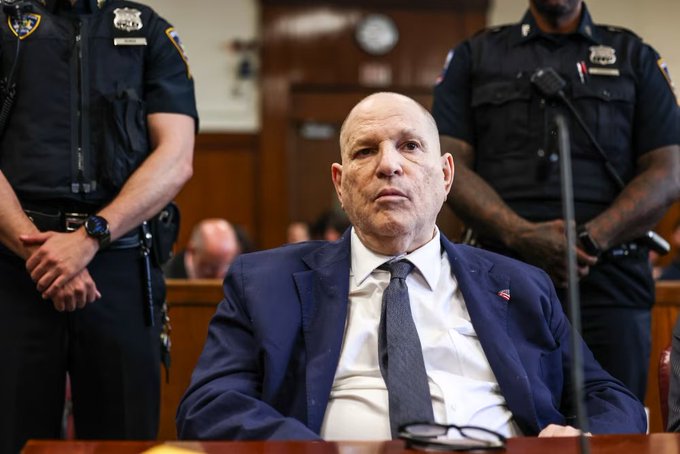 A bald man in a navy blue suit and tie sits in a courtroom setting, flanked by two uniformed police officers standing behind him.