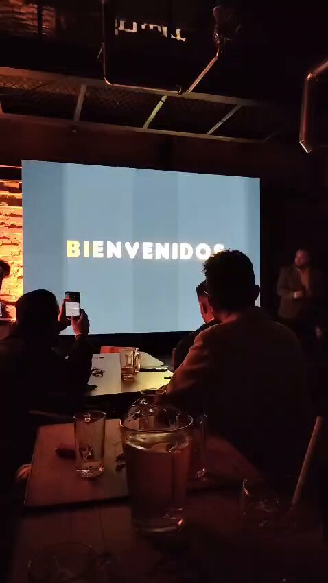 The image shows a group of people sitting around a table with drinks at a bar or restaurant. One person is taking a picture of the screen, which displays the words "bienvenidos" written in white letters. The room has a dark ambiance and features a large projection screen mounted on the wall above the dining area. There are several cups placed on the table, along with other items such as a cell phone and a bottle.