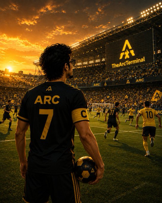 The image shows a soccer player wearing a black jersey with the number 7, holding a soccer ball and standing on a field at a stadium during an evening game. The stadium appears packed, indicating that this is a popular event or match. The lights are turned on, illuminating the scene, and there's a large screen displaying "TheARC" in yellow letters above the crowd. The player seems to be ready for action as he prepares to play against his opponents.
