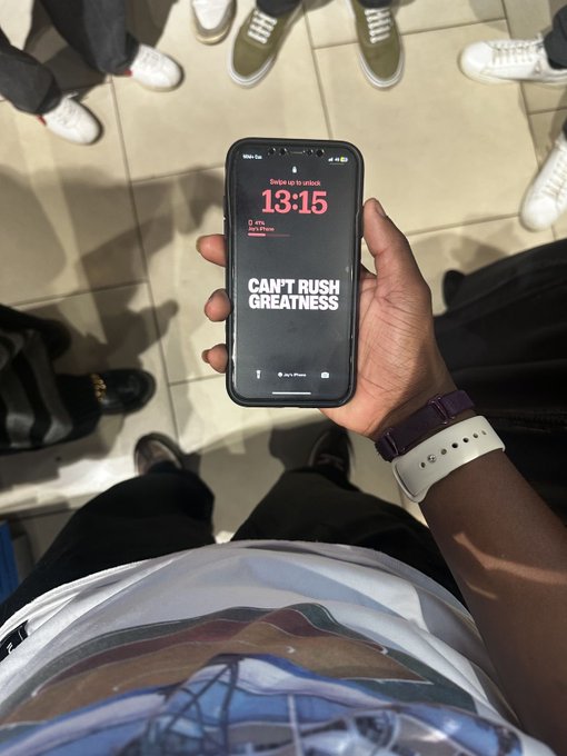 A first-person view of someone holding a smartphone displaying a lock screen that reads 'CAN'T RUSH GREATNESS' with a timer at 13:15, surrounded by other people's feet on a tiled floor.