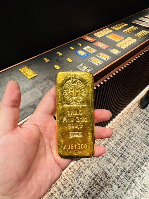 A hand holding a 1-kilo fine gold bar stamped 'ARGOR-HERAEUS SA' with serial number AJ61500, displayed in front of a counter showing additional gold bars.