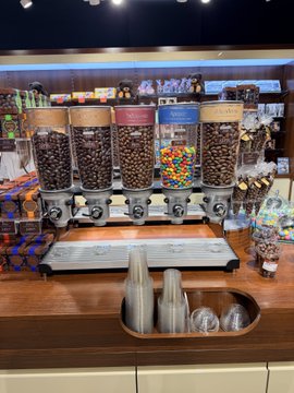 A candy/dessert shop counter featuring cereal dispensers filled with chocolate treats and colorful M&Ms, with stacks of cups in the foreground.