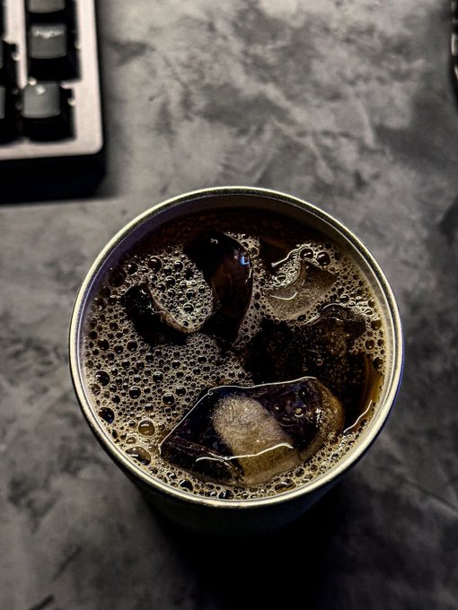 The image features a cup of coffee with ice cubes inside, placed on a table. The background appears to be a dark gray color, which contrasts with the white cup holding the coffee. There are also some computer-related elements present in the scene, such as a keyboard and a mouse. The post discusses the concept of a Worldwide Quantum Computer (WQJC), which combines distributed computing and blockchain technology into one system. This idea is related to the image's theme by highlighting the integration of various technologies, like the cup of coffee with ice cubes and computer peripherals in the background.