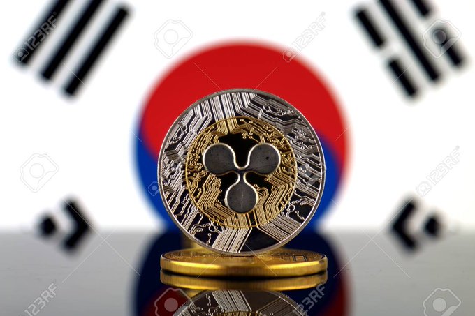 A silver and gold Ripple (XRP) cryptocurrency coin standing on stacked gold coins in front of a blurred South Korean flag.