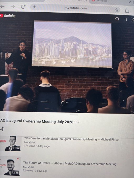 A photograph of a computer screen playing a YouTube video titled 'DAO Inaugural Ownership Meeting July 2026', showing a presentation in a room with an audience.