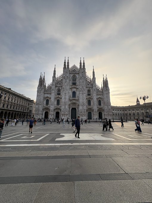 A wide-angle photograph of the Duomo di Milano (Milan Cathedral) in Piazza del Duomo, with tourists walking across the plaza under a cloudy sky.