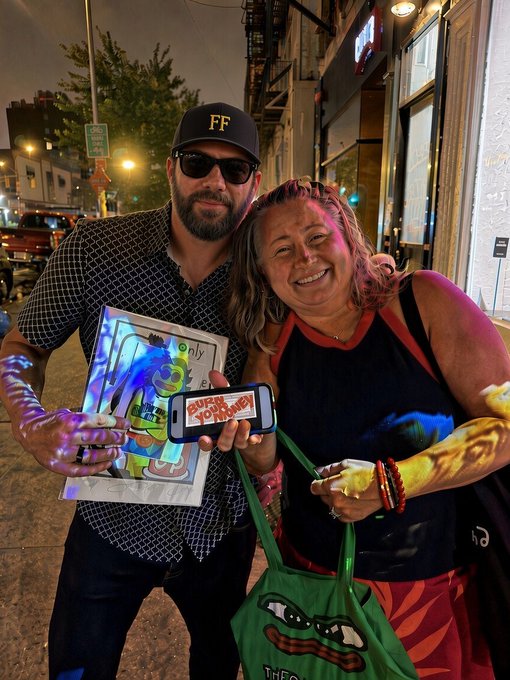 Two smiling people pose on a city street at night; the man wears an 'FF' cap and sunglasses holding artwork, while the woman holds a phone displaying 'SUPER COOL TOURNEY' and a green tote bag, with paint visible on their arms.