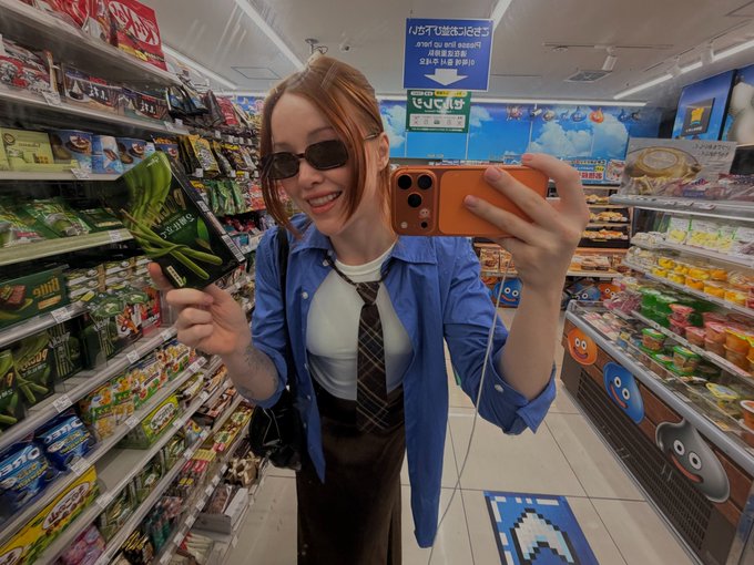 A woman wearing sunglasses and a blue shirt with a tie taking a selfie in a convenience store aisle while holding a package of green vegetables, with Chinese/Japanese signage visible in the background.