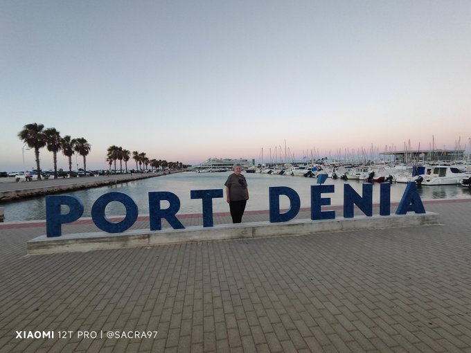 A woman stands behind large blue letters spelling 'PORT DÉNIA' on a waterfront promenade at dusk, with palm trees, a marina filled with boats, and the sea in the background; watermarked 'XIAOMI 12T PRO | @SACRA97'.