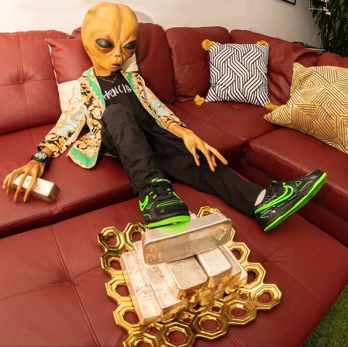 A person wearing a large alien mask, gold-patterned jacket, black Balenciaga shirt, black pants, and neon green and black sneakers lounging on a red leather couch with silver bars and gold decorative pieces on the table in front of them.